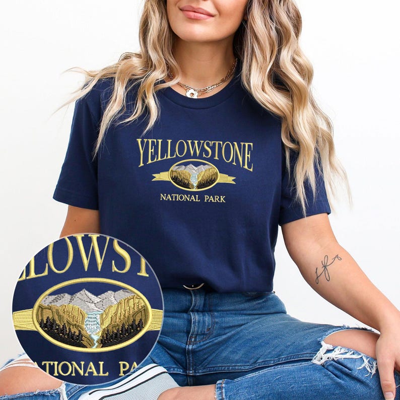 May include: Navy blue t-shirt with the embroidered text "YELLOWSTONE NATIONAL PARK" in gold. The design features a mountain and river scene within a gold oval. The shirt is a short-sleeved crew neck.