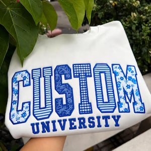 May include: A white crewneck sweatshirt featuring the word CUSTOM in large, block-style letters with varied blue and white patterns, including floral, striped, and gingham prints. The word UNIVERSITY is embroidered below in solid blue letters.