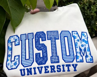 May include: A white crewneck sweatshirt featuring the word CUSTOM in large, block-style letters with varied blue and white patterns, including floral, striped, and gingham prints. The word UNIVERSITY is embroidered below in solid blue letters.