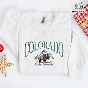May include: White sweatshirt featuring the word "COLORADO" in green script above an embroidered image of a bison and mountains. The text "Rocky Mountain" is below the bison. The sweatshirt is folded on a white surface.