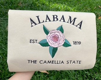 Può includere: Una felpa bianca con il testo "ALABAMA EST. 1819 THE CAMELLIA STATE" ricamato in nero. Un fiore di camelia rosa con foglie verdi è ricamato al centro.