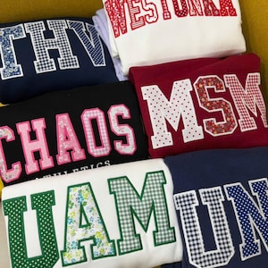 May include: A collection of folded sweatshirts in various colors, each featuring large, embroidered lettering. The letters are decorated with floral, polka dot, and gingham patterns. The sweatshirts are in white, navy, black, and burgundy.