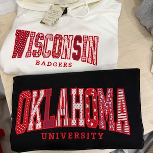 以下が含まれることがあります： 白いフード付きスウェットシャツには赤い文字でWISCONSIN BADGERS、黒いスウェットシャツには赤い文字でOKLAHOMA UNIVERSITYと書かれています。文字には水玉模様、ストライプ、花柄など、さまざまな赤と白のパターンが施されています。