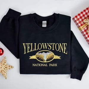 May include: Black sweatshirt with gold embroidered text "YELLOWSTONE NATIONAL PARK" and a scenic oval design. The design features mountains, trees, and a river. A gift box and Christmas ornaments are in the background.