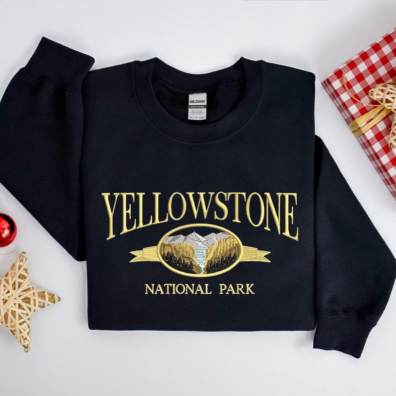 May include: Black sweatshirt with gold embroidered text "YELLOWSTONE NATIONAL PARK" and a scenic oval design. The design features mountains, trees, and a river. A gift box and Christmas ornaments are in the background.