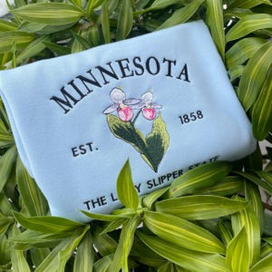 May include: Light blue sweatshirt with embroidered text "MINNESOTA EST. 1858 THE LADY SLIPPER STATE" and a pink and white flower graphic.