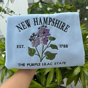 May include: Light blue sweatshirt with embroidered text "NEW HAMPSHIRE" and a purple lilac flower design. The text "EST. 1788" and "THE PURPLE LILAC STATE" are also embroidered on the sweatshirt.