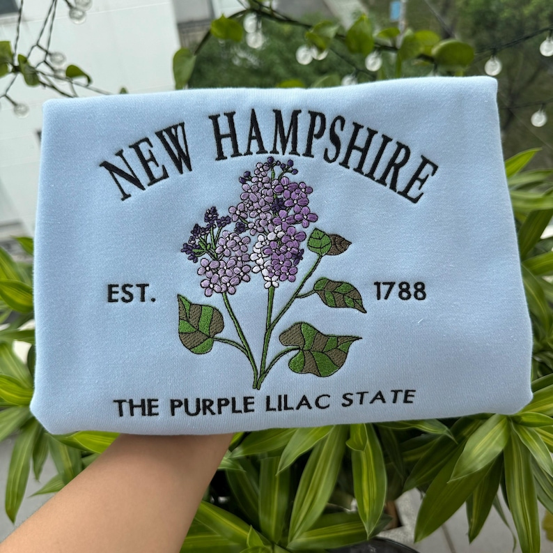 May include: Light blue sweatshirt with embroidered text "NEW HAMPSHIRE" and a purple lilac flower design. The text "EST. 1788" and "THE PURPLE LILAC STATE" are also embroidered on the sweatshirt.