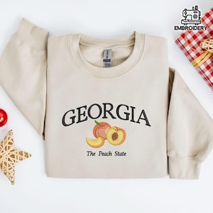 May include: A beige sweatshirt with the word "GEORGIA" embroidered in black above a peach graphic. Below the graphic, the text reads "The Peach State". The sweatshirt is folded on a white surface.