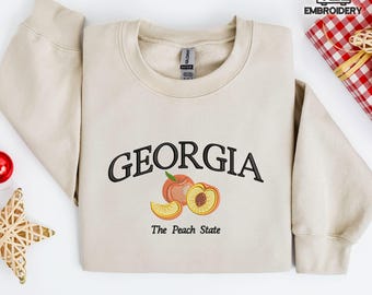 Könnte beinhalten: Beiges Sweatshirt mit dem in Schwarz gestickten Wort "GEORGIA" über einer Pfirsichgrafik. Unter der Grafik steht der Text "The Peach State". Das Sweatshirt liegt gefaltet auf einer weißen Oberfläche.