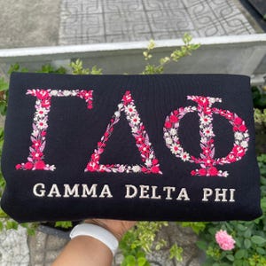 Pu&ograve; includere: Felpa nera con ricami delle lettere greche Gamma Delta Phi, decorate con motivi floreali rosa e bianchi. Il testo "GAMMA DELTA PHI" &egrave; ricamato sotto. Un capo d'abbigliamento elegante e unico.