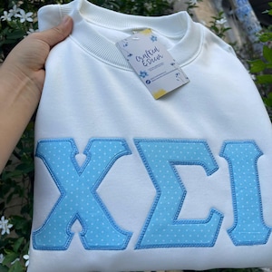 May include: A white crewneck sweatshirt featuring large, light blue Greek letters with a white polka dot pattern. The letters are outlined in matching light blue stitching. A rectangular tag labelled Crafted & Decor is attached to the collar.