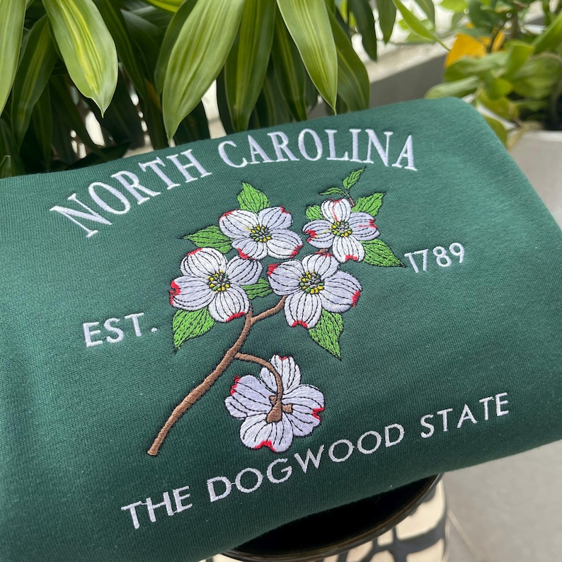 K&ouml;nnte beinhalten: Dunkelgr&uuml;nes Sweatshirt mit den Stickereien "North Carolina" und "The Dogwood State". Es zeigt ein florales Design aus wei&szlig;en Blumen mit roten Akzenten und gr&uuml;nen Bl&auml;ttern. Das Jahr "1789" ist ebenfalls aufgestickt.