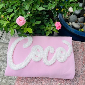May include: A pink rectangular pillow with the name "Coco" embroidered in white, fluffy script. The pillow is set against a backdrop of green foliage and pink roses, suggesting a floral theme.