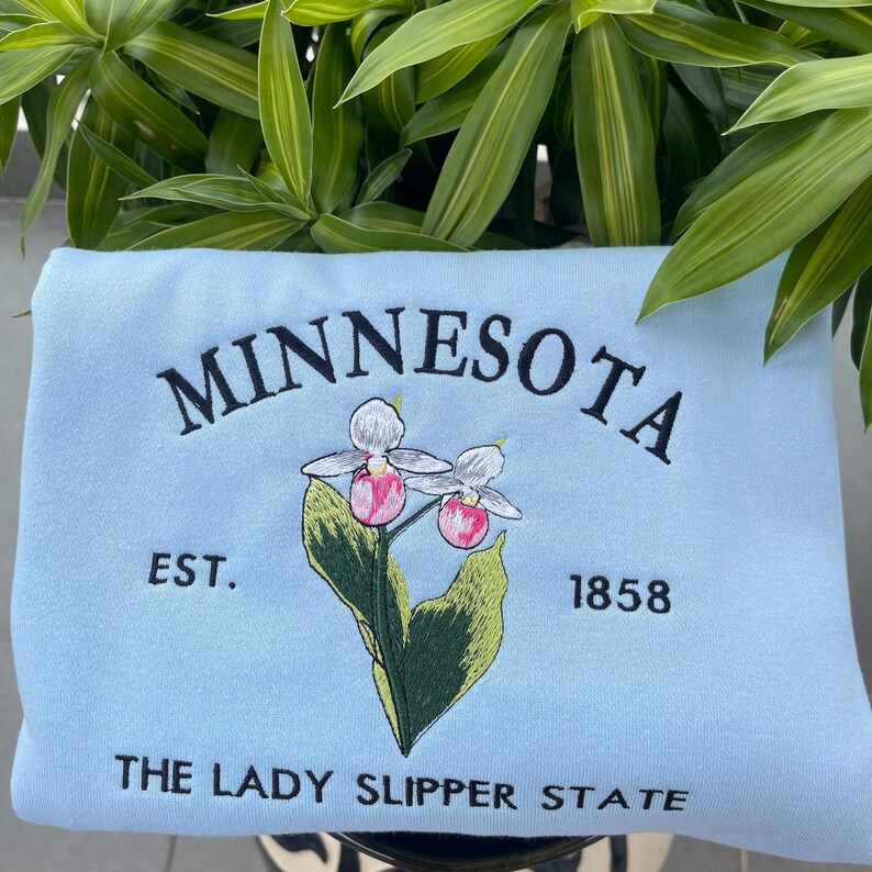 May include: Light blue sweatshirt with embroidered text "MINNESOTA EST. 1858 THE LADY SLIPPER STATE" and a stylised image of a pink and white lady slipper orchid.