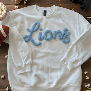 Op de afbeelding: Witte crewneck sweatshirt met het woord "Lions" in lichtblauwe, getextureerde letters over de borst. De sweatshirt heeft lange mouwen en geribde manchetten en zoom. Een voetbal en popcorn zijn zichtbaar op de achtergrond.
