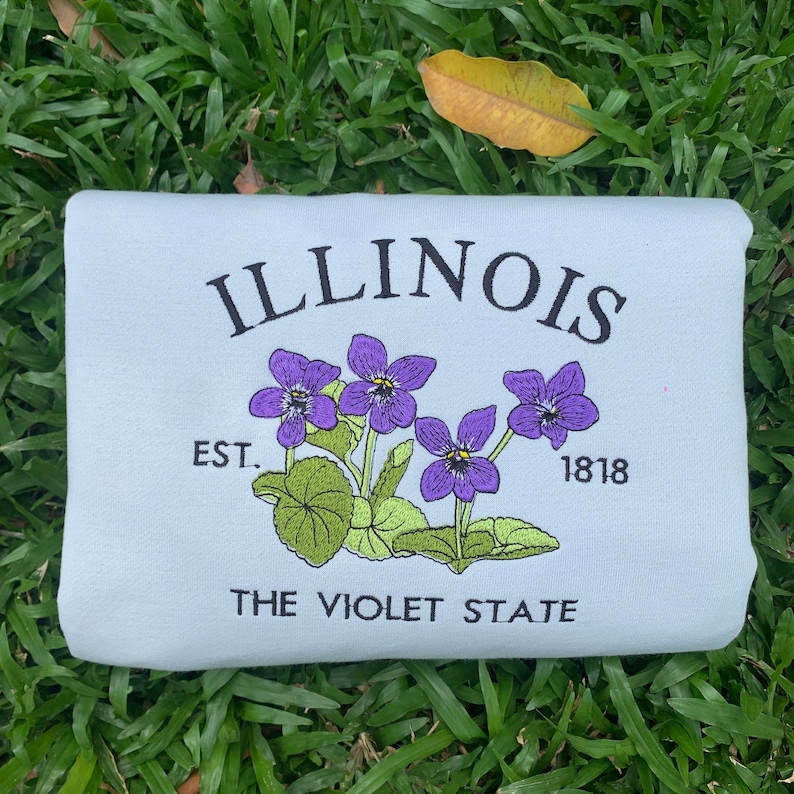 May include: Light blue sweatshirt with embroidered text "ILLINOIS EST. 1818 THE VIOLET STATE" and a purple flower design.