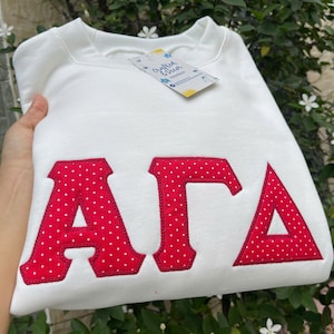 May include: A white sweatshirt with red polka-dot letters spelling out "ΑΓΑ". The letters are appliqued onto the front of the sweatshirt. A tag is attached to the neckline.