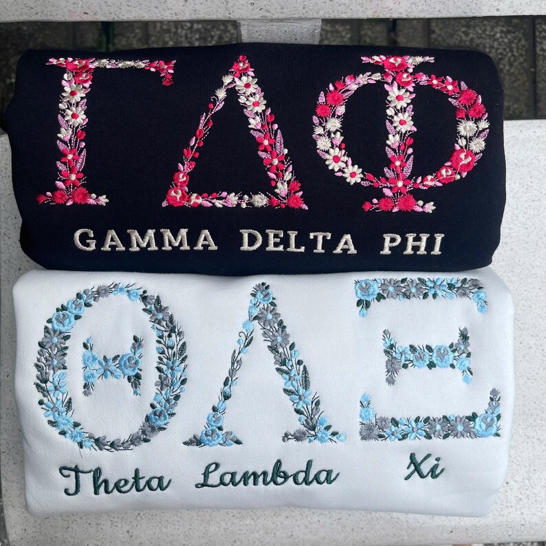 Pu&ograve; includere: Sono mostrate due felpe, una blu navy e una bianca. La felpa blu navy presenta ricami floreali che compongono "&Gamma;&Delta;&Phi;" e "GAMMA DELTA PHI". La felpa bianca ha ricami floreali che compongono "&Theta;&Lambda;&Xi;" e "Theta Lambda Xi".