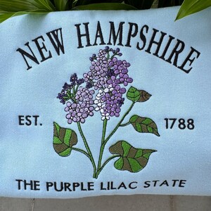 May include: A light blue sweatshirt with embroidered text that reads "NEW HAMPSHIRE EST. 1788 THE PURPLE LILAC STATE". The text is surrounded by an embroidered purple lilac flower.