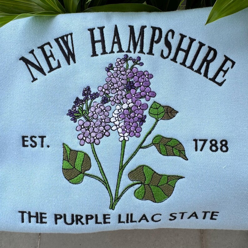May include: A light blue sweatshirt with embroidered text that reads "NEW HAMPSHIRE EST. 1788 THE PURPLE LILAC STATE". The text is surrounded by an embroidered purple lilac flower.