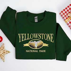 May include: A dark green sweatshirt with the embroidered text "YELLOWSTONE NATIONAL PARK". The design features a mountain and forest scene in gold and brown. The sweatshirt is folded neatly on a white surface.