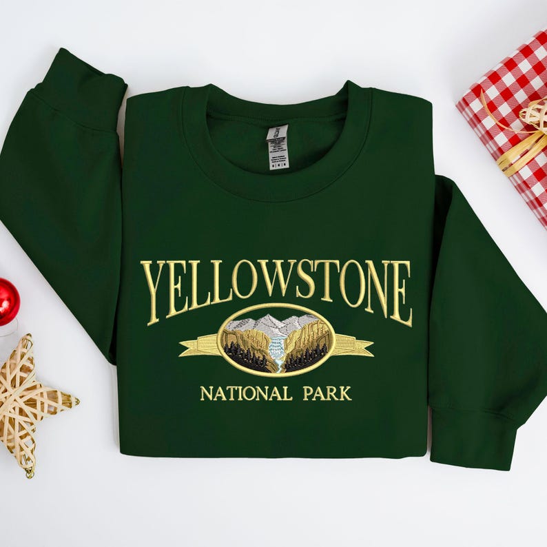 May include: A dark green sweatshirt with the embroidered text "YELLOWSTONE NATIONAL PARK". The design features a mountain and forest scene in gold and brown. The sweatshirt is folded neatly on a white surface.