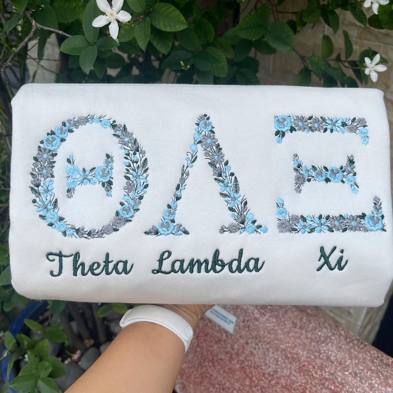 Pu&ograve; includere: Articolo rettangolare bianco con lettere greche ricamate e disegni floreali blu e grigi. Le parole "Theta Lambda Xi" sono ricamate in verde sotto.