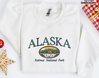Könnte beinhalten: Weißes Sweatshirt mit dem Wort "ALASKA" in Grün über einer ovalen Grafik von Bären in einer Landschaft gestickt. Unter der Grafik steht der Text "Katmai National Park". Eine rote Kugel und ein goldener Stern befinden sich in der Ecke.