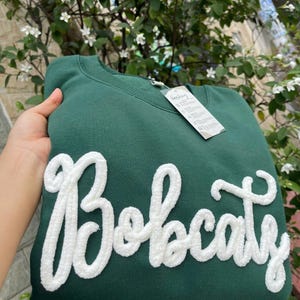 May include: A dark green crewneck sweatshirt featuring the word Bobcats in large, white, textured, fuzzy chenille-style lettering across the front. A white rectangular garment tag is attached to the neckline with printed care instructions.