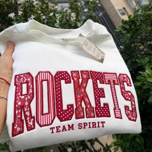 Può includere: Una felpa bianca a girocollo con la parola ROCKETS in grandi lettere applicate rosse con vari motivi floreali, a righe e a pois. Sotto la parola, il testo TEAM SPIRIT è stampato in piccole lettere maiuscole rosse.