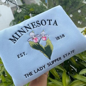 May include: Light blue sweatshirt with embroidered text "MINNESOTA EST. 1858 THE LADY SLIPPER STATE" and a pink and white flower.
