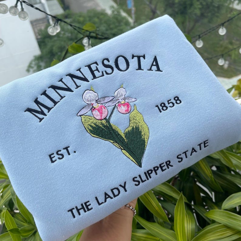 May include: Light blue sweatshirt with embroidered text "MINNESOTA EST. 1858 THE LADY SLIPPER STATE" and a pink and white flower.