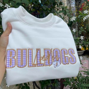 Könnte beinhalten: Ein weißes Sweatshirt mit dem Wort "BULLDOGS", das in einem lila Vichy-Muster mit goldfarbenen Umrissen gestickt ist. Unter dem Wort ist ein kleines Blumenmuster gestickt. Das Sweatshirt wird von einer Hand gehalten.