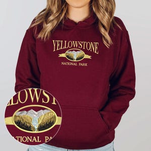 May include: A maroon hoodie with a kangaroo pocket and a drawstring hood. The front features the embroidered text "YELLOWSTONE NATIONAL PARK" in gold lettering, with a detailed mountain and river graphic in the center. The hoodie is made of a soft material.