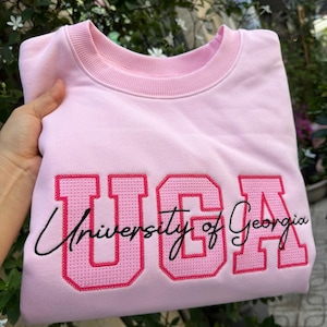 Puede incluir: Sudadera rosa claro con las letras "UGA" en rosa más oscuro, bordadas con un diseño de punto de cruz. Las palabras "University of Georgia" están escritas en cursiva negra debajo.