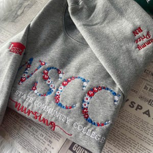 Könnte beinhalten: Graues Sweatshirt mit gesticktem Text und floralen Details. Das Design umfasst "NSCC", "NURSING" und "2024" in Rot, Weiß und Blau. Das Sweatshirt hat auch den Text "Price" und "RN" in Rot auf dem Ärmel.