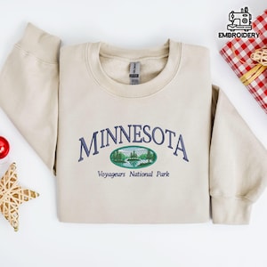 Puede incluir: Sudadera beige con la palabra "MINNESOTA" bordada en azul marino, arqueada sobre un diseño ovalado de un lago y árboles. Debajo del diseño está el texto "Voyageurs National Park". La sudadera está doblada sobre una superficie blanca.