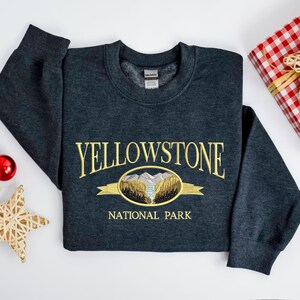 May include: A dark gray sweatshirt with the embroidered text "YELLOWSTONE NATIONAL PARK" in gold. The design includes an oval graphic with a mountain and river scene. The sweatshirt is folded on a white surface, with a red ornament and a star-shaped decoration.