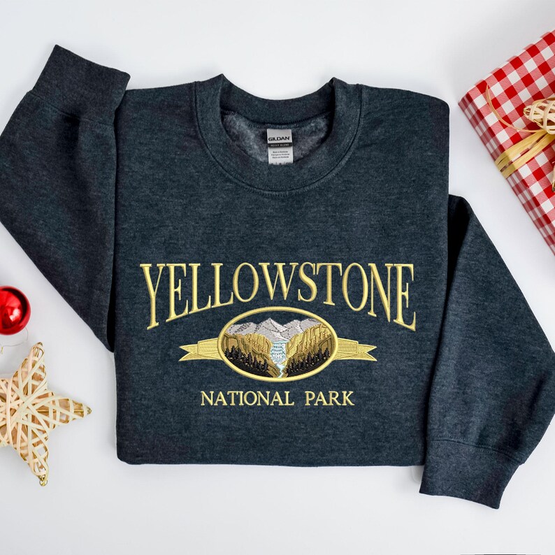 May include: A dark gray sweatshirt with the embroidered text "YELLOWSTONE NATIONAL PARK" in gold. The design includes an oval graphic with a mountain and river scene. The sweatshirt is folded on a white surface, with a red ornament and a star-shaped decoration.