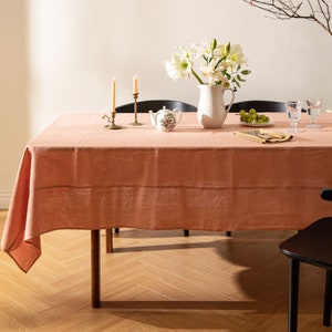 Op de afbeelding: Een lichtroze linnen tafelkleed met een subtiele textuur, gelegd op een houten tafel met een visgraatpatroon. De tafel is gedecoreerd met een witte kan gevuld met witte bloemen, een klein theepotje, twee kandelaars met kaarsen en een bord met groene druiven.