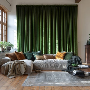 Puede incluir: Un sofá modular grande con tapicería de tela gris claro y múltiples almohadas en varios colores y texturas. El sofá está situado frente a una ventana con cortinas de terciopelo verde oscuro. Una alfombra gris cubre el suelo de madera.