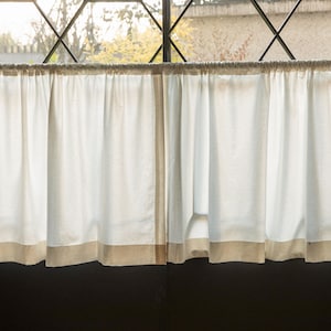 May include: Two white cafe curtains with a beige bottom border hang in front of a window with black metal bars. The curtains are pulled to the side, revealing a view of trees and a building.