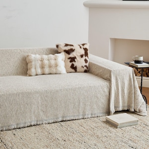 May include: A beige and white striped sofa cover with a fringed edge, featuring a rectangular cream-colored textured pillow and a square brown and white cow-print faux fur pillow placed on top of the fabric covering the sofa.