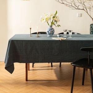 以下が含まれることがあります： 濃いグレーのリネン製のテーブルクロスが、木製のテーブルにかけられています。テーブルの上には、青と白の磁器の花瓶に白い花が活けられています。また、真鍮製の燭台に白いろうそくが2本、透明なガラスのワイングラスが2つ置かれています。