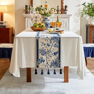 May include: White tablecloth with a blue and white floral patterned table runner with tassels. The table runner has a blue border. The table is set with a vase of flowers, fruit, and two wine glasses.