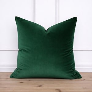 May include: A square, emerald green velvet pillow. The pillow is a solid color and has a soft, plush appearance. The pillow is resting on a light brown wooden surface, with a white wall in the background.