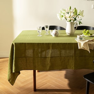 May include: A green linen tablecloth is laid out on a wooden table with a teapot, two glasses, a plate of grapes, and a napkin. White flowers in a pitcher are on the table.