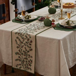 Pode incluir: Uma mesa de jantar festiva com uma toalha de mesa creme e um caminho de mesa com um design botânico verde e vermelho. Os individuais e guardanapos verdes complementam o caminho. Taças de champanhe e abóboras decorativas adicionam ao ambiente festivo.