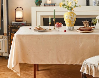 Puede incluir: Un mantel beige con un sutil patrón de bambú, preparado para una cena con un jarrón de flores blancas, velas y un cuenco de fruta. El mantel está puesto sobre una mesa de madera con una silla en el fondo.